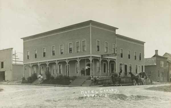 Photographic postcard view of the Hotel Carley. Two women are sitting on the far left steps of the hotel. One man is sitting in a chair near the entrance, with a man standing to his left. Three youths are standing nearby looking towards a man on a horse, and another man is sitting and holding the reins of a horse-drawn wagon. Caption reads: "Hotel Carley, Aniwa, Wis."