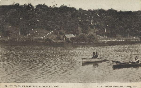 Photographic postcard view across water towards a tree-covered shoreline near Dr. Whitcomb's Sanitarium. Two men are sitting in a rowboat near another man in a rowboat in the foreground. Caption reads: "Dr. Whitcomb's Sanitarium, Albany, Wis."