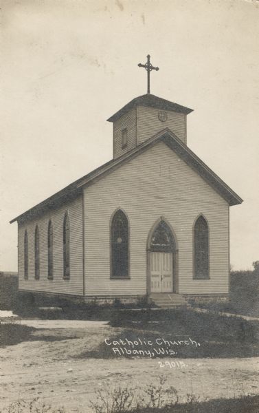Photographic postcard view of a white, one-story Catholic Church. Exterior view of the main entrance, including stained glass Gothic-Revival styled windows above the entrance and along the side of the building. There is a cross above the church's steeple. Caption reads: "Catholic Church, Albany, Wis."