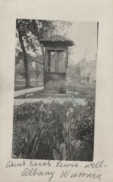 Photographic postcard view of an old wooden well. Tulips line the sidewalk near the well. Residential houses, trees and yards are in the background.