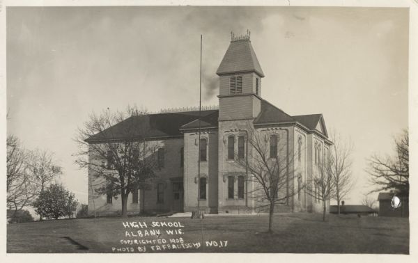 Photographic postcard view of the exterior of the high school. Front view of the school includes a flag pole and bare trees on the lawn surrounding the school. Several other buildings are in the background. Caption reads: "High School, Albany, Wis."
