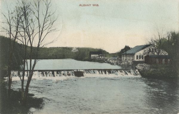 Elevated view toward the dam. The shoreline is lined with trees, and there are buildings near the dam on the right Caption reads: "Albany, Wis."