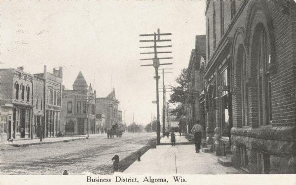 Photographic postcard of the business district. Sidewalk view shows pedestrians and a vehicle in the street. Telephone poles and small horsehead shaped hitching posts along the sidewalk.