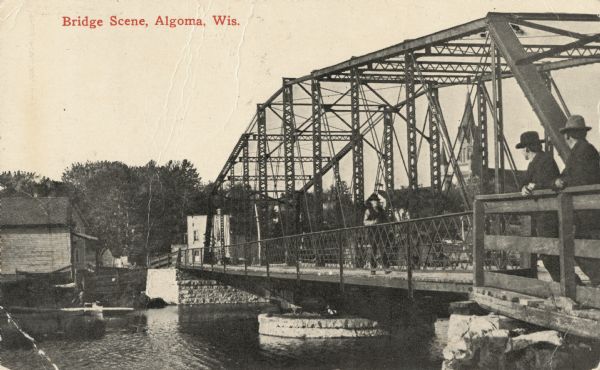Photographic postcard shoreline view of a bridge. Two men in hats are standing on the far right at a railing on the truss bridge looking toward the opposite shore, and another man is walking towards them. The opposite shoreline and buildings are on the left, and a church steeple is behind the bridge in the far background. Caption reads: "Bridge Scene, Algoma, Wis."