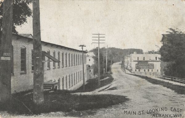 View of Main Street looking east. Trees and telephone poles line the street next to a large two-story building. A bridge and sidewalk are in the middle-ground and a grocery/clothing house and other businesses are visible on the opposite side. Caption reads: "Main St. Looking East, Albany, Wis."