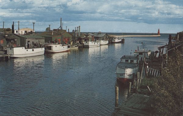 Color photographic postcard of the fisherman's dock taken from the harbor walkway. White boats are moored on either side of the Ahnapee River as it flows into Lake Michigan. The red Algoma Pierhead Lighthouse is at the end of the northern pier. The first pierhead lighthouse was established in 1893 and replaced in 1908 by a cast-iron tower, which was mounted on top of a cylinder in 1932 to raise the focal point.