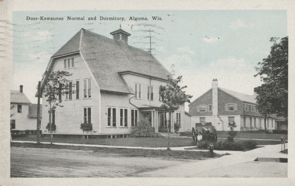 Colorized postcard view of the exterior and lawns of the Door-Kewaunee Normal School and its dormitory. Street view of the school with  a few small trees lining the street. The dormitory is in the background to the right of the school. A young boy is standing in front of a cannon.