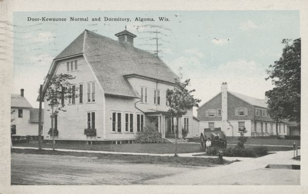 Colorized postcard view of the exterior and lawns of the Door-Kewaunee Normal School and its dormitory. Street view of the school with  a few small trees lining the street. The dormitory is in the background to the right of the school. A young boy is standing in front of a cannon. Caption reads: "Door-Kewaunee Normal and Dormitory, Algoma, Wis."