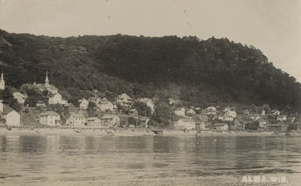 Photographic postcard from the Mississippi of the city of Alma. The city of Alma is along Wisconsin State Highway 35, also known as the Great River Road. The rocky, tree-covered bluffs rise up behind the city. Caption reads: "Alma, Wis."