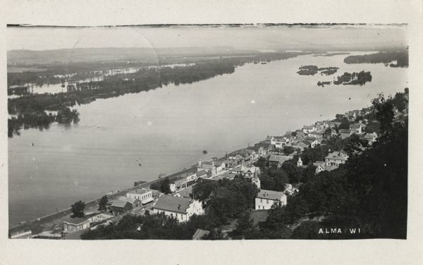 Photographic postcard of elevated view of the city and the Mississippi River. Homes and business are visible along 2nd Street. Caption reads: "Alma, WI."