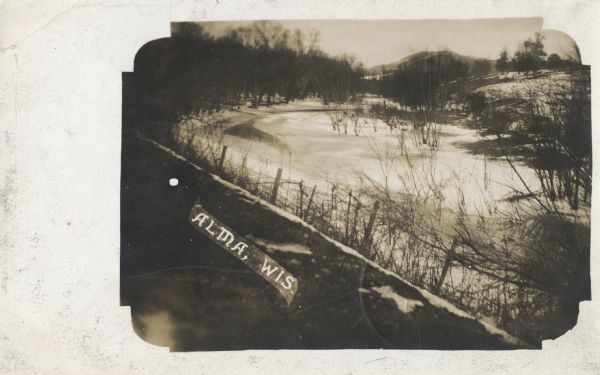 Photographic postcard of a frozen body of water. Ice and snow mostly cover a pond or river, with some open water. Bare trees and a barbed wire fence are in the foreground. Caption reads: "Alma, Wis."