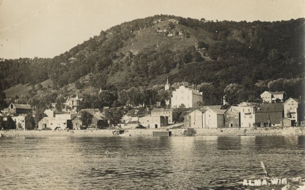 Photographic postcard view of southern Alma with buildings and bluffs. The Mississippi River and the shoreline are in the foreground. Caption reads: "Alma, Wis."
