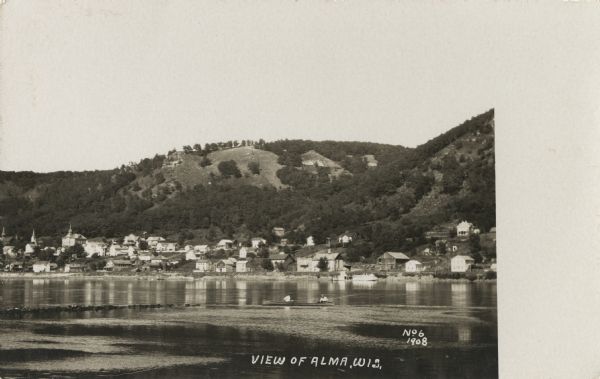 Photographic postcard view over the Mississippi River showing of the town of Alma. Bluffs are behind the town. In the foreground, two men wearing hats are sitting in a canoe with fishing rods. Caption reads: "View of Alma, Wis."