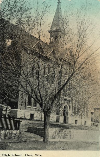 Colorized photographic postcard view of the exterior of the high school, which has  two-stories, and is surrounded by a lawn. A tree is obscuring the facade and bell tower of the school. Caption reads: "High School, Alma, Wis."