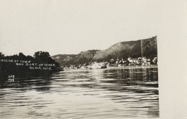 Scene of the town of Alma from the Mississippi River. Alma and the bluffs along the Mississippi are in the far background. A paddle steamer can be seen floating down the river past the town. Caption reads: "Scene of Town and Boat Up River, Alma, Wis."