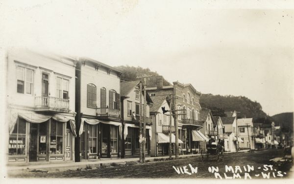 View across street of shops and other buildings along Main Street. A buggy is driving down the road. There is a bluff in the background. Caption reads: "View on Main St. Alma, Wis."