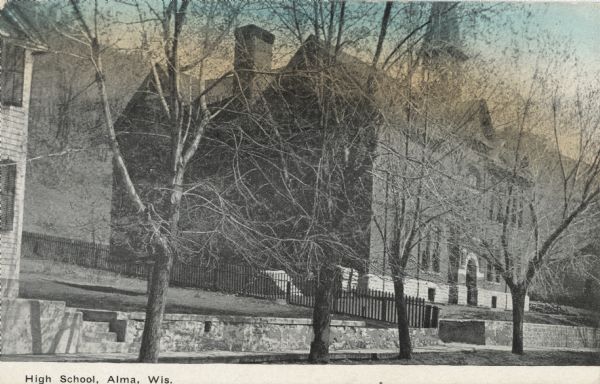 Colorized photographic postcard from across a street of the high school. There is a stone wall in front of the school, and a steep hill behind. Caption reads: "High School, Alma, Wis."