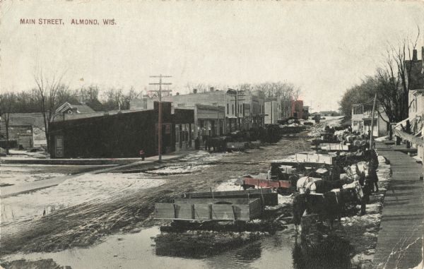 Elevated view down Main Street. Horse-drawn wagons line both sides of the road which is covered with puddles, mud and snow. Caption reads: "Main Street, Almond, Wis."