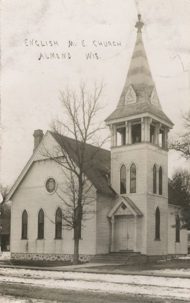Photographic postcard of the exterior of the one-story, white English Methodist Church with belfry and steeple. Snow covers the street outside the church. Caption reads: "English M.E. Church, Almond, Wis."
