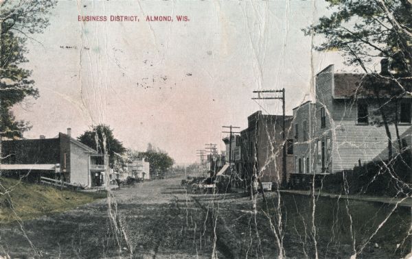 Color photographic postcard of the business district. Local business and homes are on on both sides of the main road. Horse-drawn wagons are parked on the right. Caption reads: "Business District, Almond, Wis."C