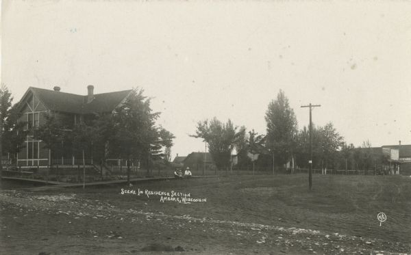 Photographic postcard view across dirt road toward two men and a child sitting on the sidewalk in front of a house in residential area. Caption reads: "Scene in Residence Section, Marinette, Wis."