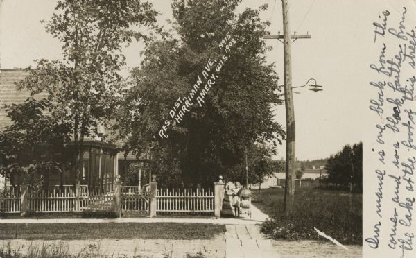 Photographic postcard view of Harriman Avenue in the residential district. A little girl is standing with her baby carriage on the sidewalk between a telephone pole and a house. Caption reads: "Res. Dist. Harriman Ave., Amery, Wis."