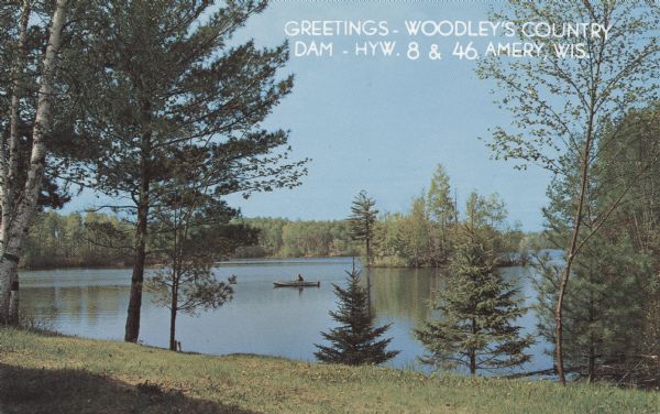 Color photographic postcard of a man in a fishing boat on Woodley's Dam north of Amery. View of the dammed water surrounded by pine trees. Caption reads: "Greetings - Woodley's Country Dam-HWY.8 & 46 Amery, Wis."