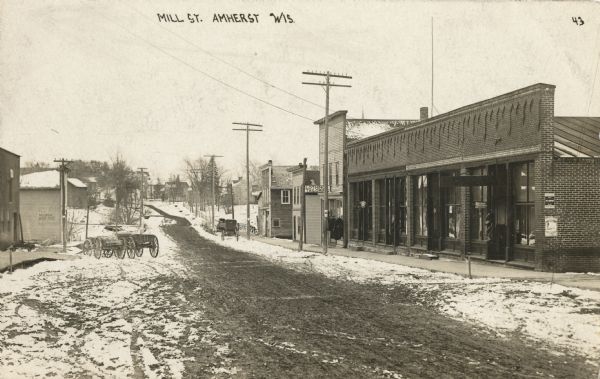 Photographic postcard of Mill Street. Town businesses, including a clothing and tailoring store, line the right side of the street looking towards a residential neighborhood. Snow-covered wagons are parked along the curb. aption reads: "Mill Street, Amherst, Wis."
