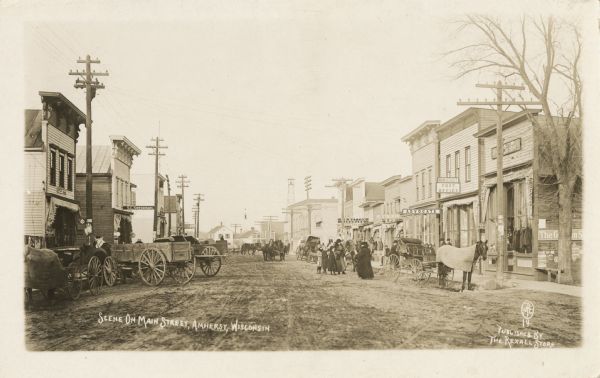 Photographic postcard looking down Main Street. Horse-drawn wagons and people line the street next to local businesses, including the post office and hardware store. Caption reads: "Scene on Main Street, Amherst, Wis."