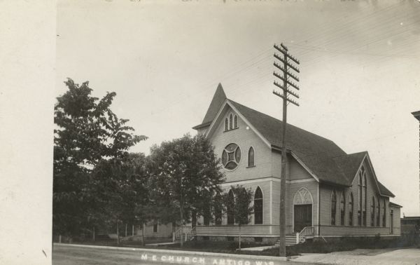 Photographic postcard view across street toward the church. The church's bell tower is in the background. Caption reads: "M.E. Church, Antigo, Wis."