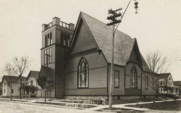Photographic postcard view of the church. The church structure includes a two-level, square belfry on the left. Two small children are sitting on a four-wheeled wagon on the corner at the sidewalk. Caption reads: "Congregational Church, Antigo, Wis."