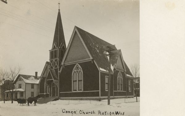 Photographic postcard view of the church. The church structure includes a two-level, square belfry with a pointed steeple. A horse-drawn sled is sitting outside along the snow-covered street. Caption reads: "Cong. Church, Antigo, Wis."