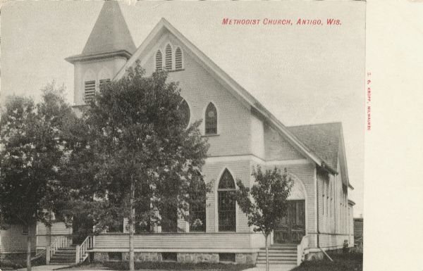 Photographic postcard view church. Trees partly obscure the front facade. Caption reads: "Methodist Church, Antigo, Wis."