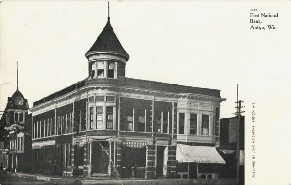 Photographic postcard view of the entrance to the First National Bank. The two-story building with turret stands among other buildings in the downtown area. Caption reads: "First National Bank, Antigo, Wis."