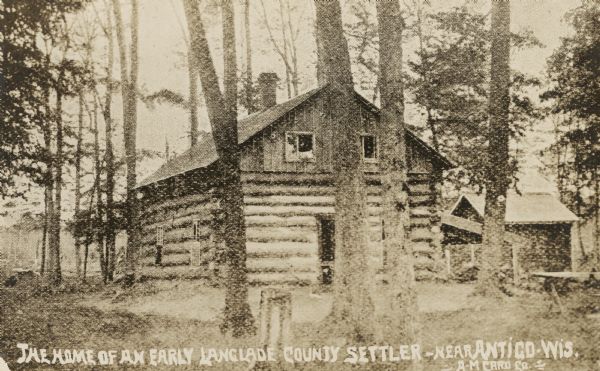 Two-story log cabin and shed in a forest. The building was used by early settlers in Langlade County near Antigo. Caption reads: "The Home of an Early Landglade County Settler — near Antigo, Wis."