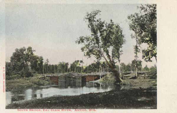 View of the shoreline and South Bridge suspended over the Eau Claire River. Caption reads: "South Bridge, Eau Claire River, Antigo, Wis."