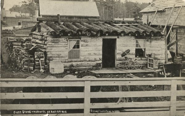 Photographic postcard of the exterior of the first store and hotel in Antigo. A baby carriage is sitting outside the front door to the building, and a pile of cut wood is piled on the side. A fence is in the foreground and other buildings are in the background. Caption reads: "First Store and Hotel at Antigo, Wis."