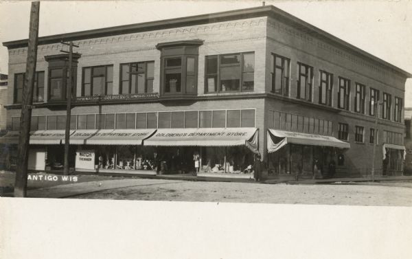 Photographic postcard view across intersectiontoward the exterior of the two-story Goldberg's Department Store. Various goods are on display in the storefront windows, and an overhang displays the name of the store. Caption reads: "Antigo, Wis."