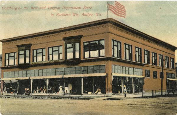 Exterior of Goldberg's Department Store. Merchandise is displayed in the storefront windows and an American flag is on the roof of the building. Caption reads: "Goldberg's — The Best and Largest Department Store in Northern Wis. Antigo, Wis."