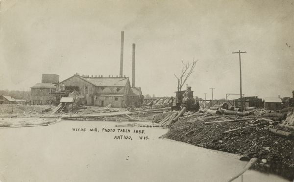 Photographic postcard view of Weeds Mill. The mill has smokestacks and logs piled on the pond shoreline waiting for processing. A steam locomotive is on the right with flatbeds of logs piled in stacks. Caption reads: "Weeds Mill, Photo Taken 1885, Antigo, Wis."