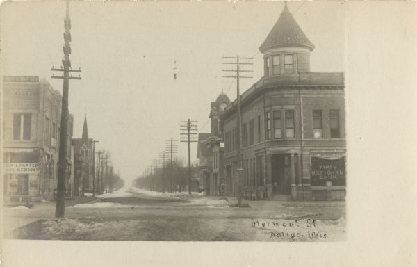 Photographic postcard view of downtown looking down Clermont Street. The two-story First National Bank is on the right among other businesses and a church building. Small piles of snow are along the street. Caption reads: "Clermont Street, Antigo, Wis."