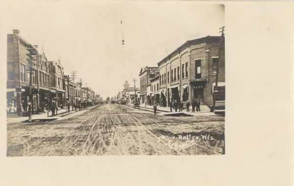 Photographic postcard view of 5th Avenue. Citizens and horse-drawn wagons line the streets and sidewalks. Stores are on both sides of the street, and a bank is at the intersection on the right. Caption reads: "5th Avenue, Antigo, Wis."