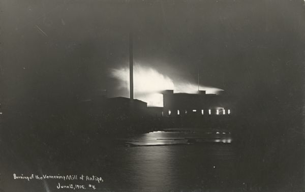 Photographic postcard of a night view over water of the veneering mill at Antigo burning. Flames and smoke are streaming from the roof and windows of the building. Caption reads: "Burning of the Veneering Mill at Antigo."