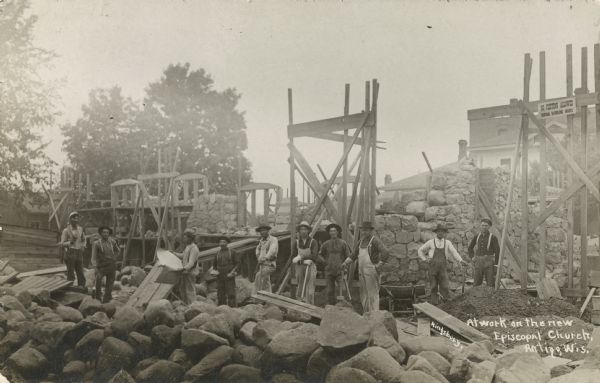 Photographic postcard view of workers building the new Episcopal Church. Workers in hats and overalls, and holding shovels, are standing among scaffolding, rocks, and other construction equipment. Caption reads: "At Work on the New Episcopal Church, Antigo, Wis."