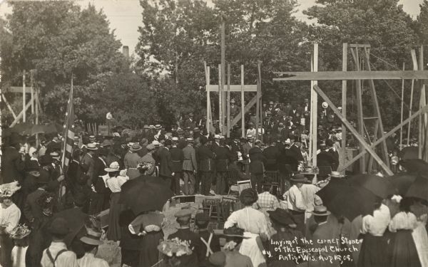 Photographic postcard of the crowd gathered to see the laying of the corner stone for the new Episcopal Church. Men and women, many of them wearing hats, are standing among scaffolding at the construction site. Caption reads: "Laying of the Corner Stone of the Episcopal Church, Antigo, Wis."
