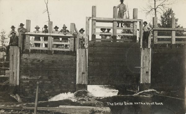 Photographic postcard view looking up at twelve workers wearing hats posing on top of the Dells Dam on the Wolf River. Water is flowing through the dam at the bottom. Caption reads: "The Dells Dam on the Wolf River."
