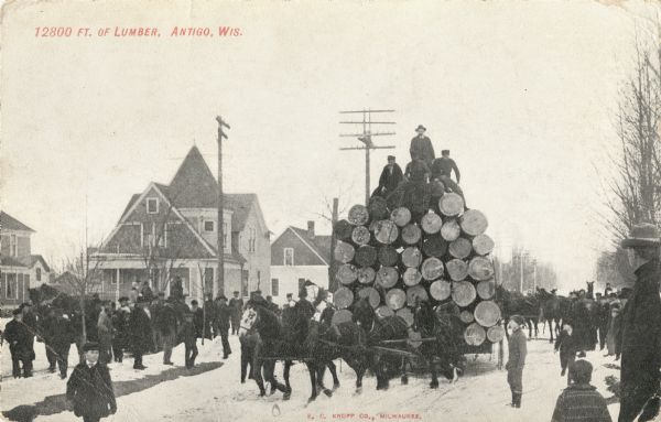 Team of horses driving through Antigo hauling 12800 Ft. of lumber. Three men and one driver are sitting on top of the large pile of wood as a crowd of people are watching as the team drives through the snow-covered street. Caption reads: "12800 Ft. of Lumber, Antigo, Wis."