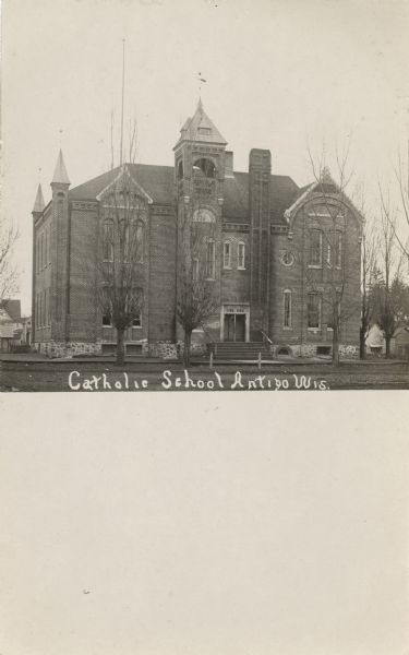 Exterior view of the front entrance to the two-story building. Bare trees are standing in the lawn. Caption reads: "Catholic School, Antigo, Wis."