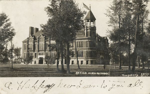 Photographic postcard view of the Antigo high school. Trees with leaves cover the lawn, and an American flag is flying on the roof of the building behind the bell tower. Caption reads: "Antigo High School, Antigo, Wis."