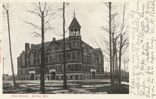 Three-quarter view of the high school which has a bell tower. Bare trees and piles of snow cover the lawn. Caption reads: "High School, Antigo, Wis."